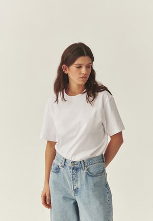 Young woman with long dark hair wearing a white t-shirt and light blue high-waisted jeans, looking down and to the side against a plain background.