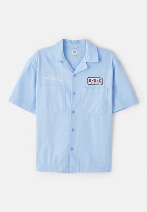 Hellblaues Kurzarmhemd aus Baumwolle mit zwei Fronttaschen, Knopfleiste und einem Aufnäher-Logo mit roten und marineblauen Akzenten.