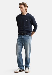 Marineblå sweatshirt med hvid krave, lyseblå jeans og hvide sneakers med sorte detaljer, stående positur mod en hvid baggrund.