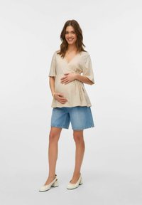 Gai bēšs apvalks ar īsām piedurknēm, apvienots ar ziliem džinsu šortiem. Modelis stāv ar rokām uz vēdera, ģērbies baltās blokveida papēžos.