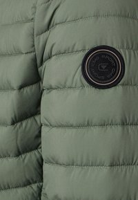 Grön vadderad jacka med quiltat mönster, som har en rund svart logotypapplikation på den övre ärmen och kontrasterande sömmar.