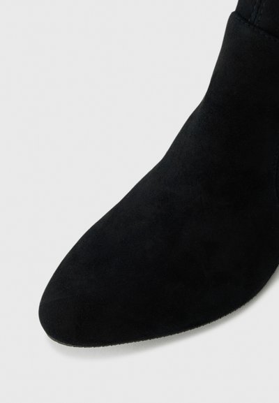 Prim-plan al unei cizme de gleznă din suede negru, cu vârf rotund, pe un fundal deschis.