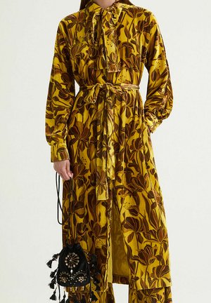 Longue robe jaune avec des imprimés floraux marron, un col à nouer et une ceinture assortie. Comprend un petit sac à cordon noir avec des embellissements.