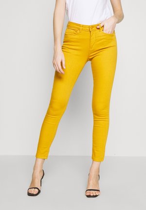 Jeans skinny amarillos con una textura suave, de cintura alta y diseño clásico de cinco bolsillos. Combinados con una blusa blanca y sandalias negras.