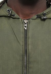 Χακί πράσινο μπουφάν με κουκούλα, διαθέτει μεταλλικό φερμουάρ πλήρους μήκους, κορδόνια στη κουκούλα και απαλό, ελαφρώς ανακλαστικό υλικό.