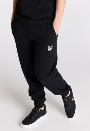 Pantalones de chándal negros con bolsillos laterales, con un logo blanco en el muslo izquierdo. Combinados con zapatillas negras con detalles dorados y suela blanca.