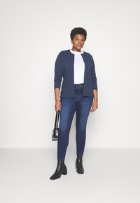 Navy ribbestrikket cardigan over en hvit topp, kombinert med mørkeblå skinnyjeans og svarte ankelstøvler. Holder en liten svart håndveske.