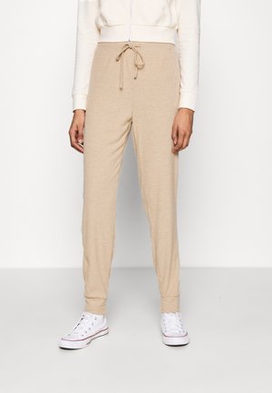 Beige geribbelde gebreide broek met trekkoord in de taille, gedragen met een witte jas met rits en witte hoge sneakers op een lichte achtergrond.
