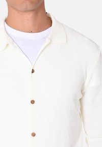 Cardigan lavorato a maglia color crema con colletto, bottoni in legno e tessuto texturizzato. Indossato su una camicia bianca.