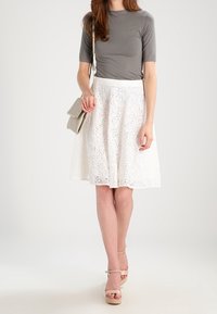 Graues eng anliegendes Oberteil kombiniert mit einem weißen, strukturierten knielangen Rock mit Blumenmuster. Das Model trägt eine hellgraue Handtasche und beige Sandalen.