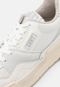 White sneaker with perforated leather and suede accents. Features a textured beige sole and a label with "GANT" in gray on the tongue.