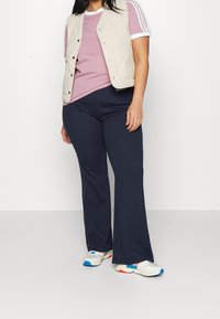 Kvinna i rosa kortärmad skjorta med vita ränder, beige vadderad väst, svarta utsvängda byxor och vita sneakers med blåa och röda detaljer.