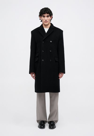 Un jeune homme se tenant debout, vêtu d'un manteau noir croisé, de pantalons gris à large jambe et de chaussures noires, sur un fond blanc uni.