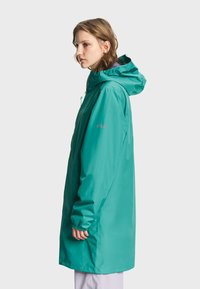 Jeune personne portant un imperméable à capuche bleu sarcelle, debout de profil avec les mains détendues le long du corps sur un fond uni.