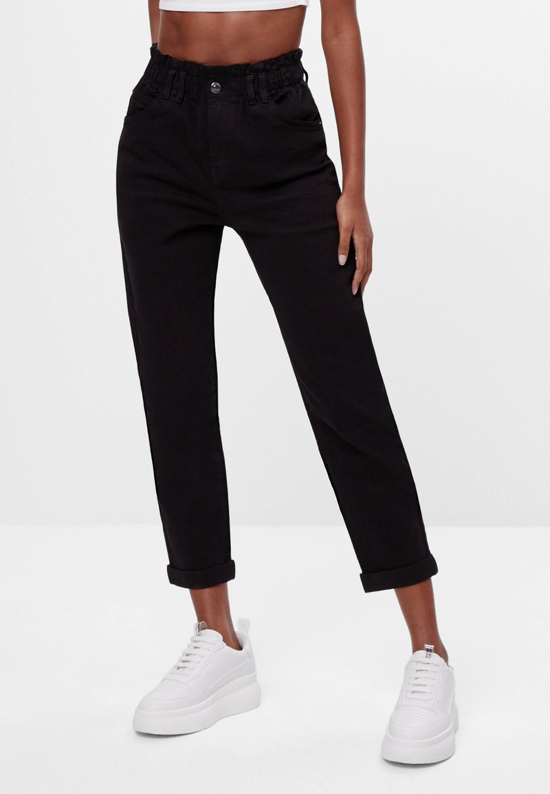 Pantalons noirs taille haute avec une ceinture élastique à volants, jambes droites et ourlets retroussés. Assortis à des baskets blanches.