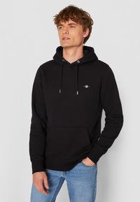 Melns džemperis ar kapuci no mīksta auduma, ar priekšējo kabatu, auklām un nelielu izšūtu logotipu krūšu rajonā. Garas piedurknes.