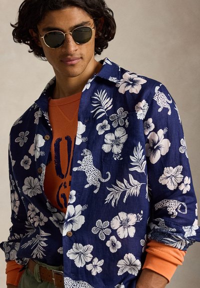 Jeune homme portant des lunettes de soleil rondes, une chemise bleu marine à motifs floraux et léopard par-dessus un sweat-shirt orange.
