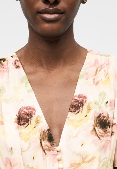 Άτομο με σκουρόχρωμο δέρμα που φοράει ανοιχτό φλοράλ φόρεμα με βαθύ V και τρία μικρά κουμπιά στο κέντρο.