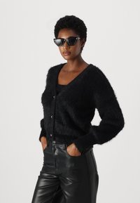 Cardigan nero peloso con scollatura a V profonda, apertura frontale con bottoni e maniche lunghe, abbinato a pantaloni in pelle neri. Gli accessori includono occhiali da sole a forma di occhio di gatto.