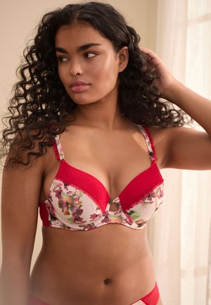 Femme aux cheveux bouclés ajustant sa coiffure, portant un soutien-gorge rouge à motif floral, debout dans une pièce doucement éclairée avec des rideaux transparents.