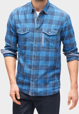 Homme portant une chemise à carreaux bleue et noire boutonnée avec les manches retroussées et un jean foncé, debout les bras détendus le long du corps.