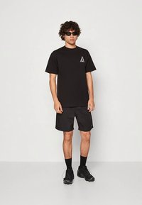 Black cotton t-shirt with short sleeves and a small triangular logo on the chest, paired with matching black shorts and sneakers.