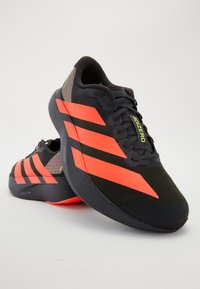 Chaussures de sport noires avec trois rayures orange audacieuses. Présentent un design épuré, une tige en maille et une semelle texturée avec un accent sur le talon bien marqué.