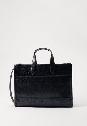 Czarna torba typu tote ze skóry, z podwójnymi uchwytami i odpinanym paskiem na ramię, z wytłoczonym napisem "MICHAEL KORS" na dolnej krawędzi z przodu.