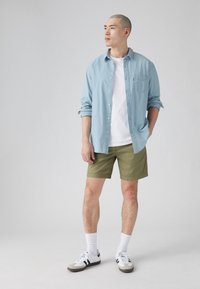 Cămașă bleu deschis cu butoni, tricou alb, pantaloni scurți verzi măslinii și adidași albi cu accente negre. Modelul stă pe un fundal neutru.