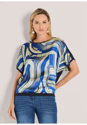 Frau mit glatten blonden Haaren trägt eine blaue und cremefarbene Bluse mit abstraktem Muster und blaue Jeans, posiert mit der Hand in der Hüfte vor beigem Hintergrund.