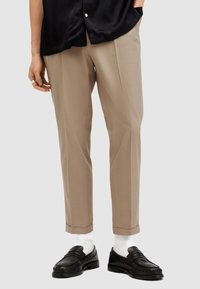 Pantaloni beige su misura con taglio dritto, tessuto liscio e orlatura, abbinati a mocassini neri e calzini bianchi alla caviglia.