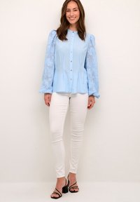 Light blue button-up blouse with sheer, embroidered long sleeves, paired with white skinny pants and black heeled sandals.