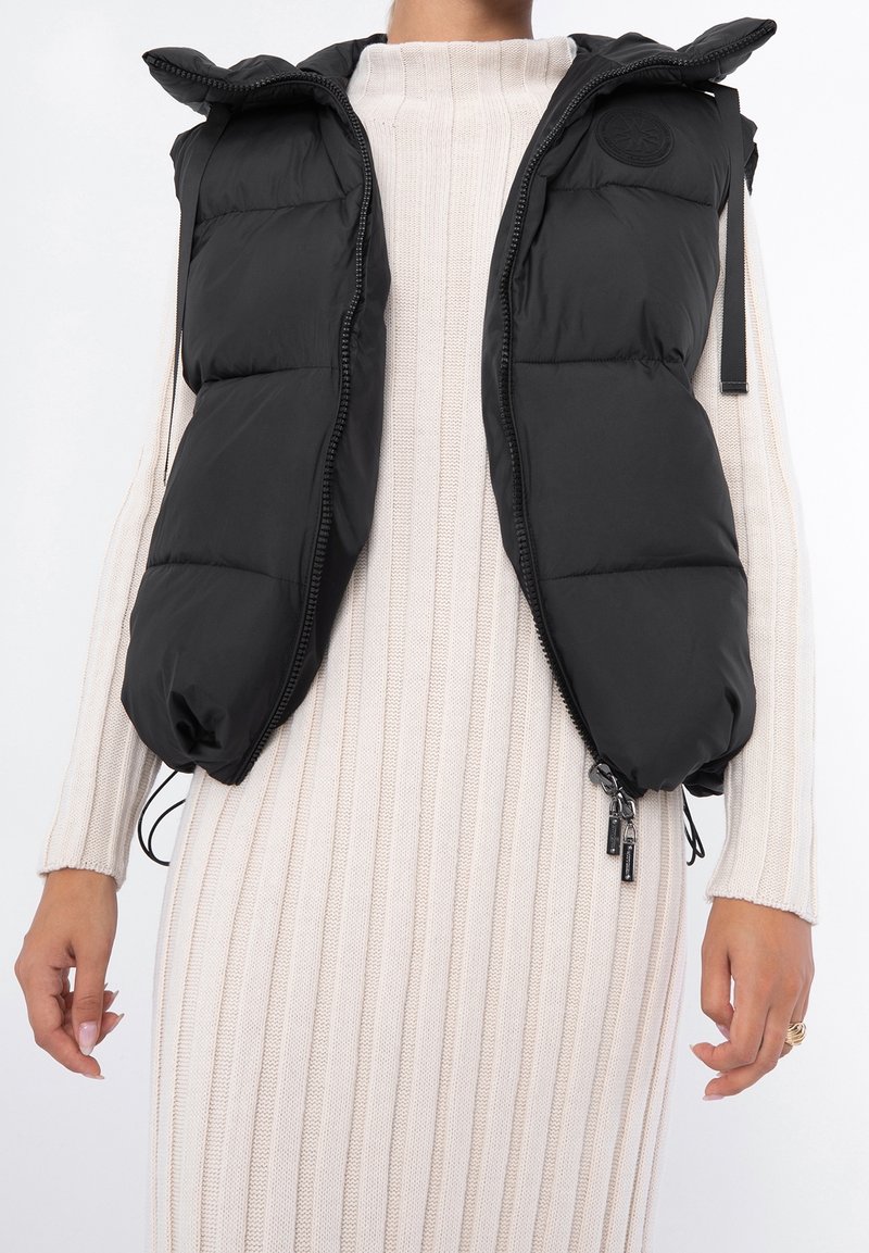 Veste matelassée noire avec fermeture éclair et capuche portée sur une robe tricotée blanche à manches longues côtelée, montrant le haut du corps et les mains.