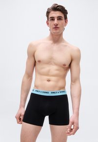 Γυμνασμένος νέος άνδρας με μαύρα ανδρικά εσώρουχα boxer με μπλε μέση που φέρει την ετικέτα "ONLY & SONS" σε απλό λευκό φόντο.