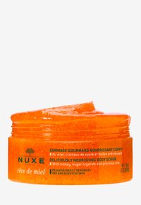 Ljust orange kroppsskrubb i en klar burk, texturerad med sockerkrystaller. Etiketten visar produktnamn och viktiga ingredienser på flera språk.