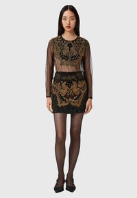 Schwarzes, durchsichtiges Oberteil mit goldenen Verzierungen, taillierter schwarzer Rock, lange Ärmel und ein strukturiertes Muster. Das Model trägt durchsichtige Strumpfhosen und schwarze Schuhe.