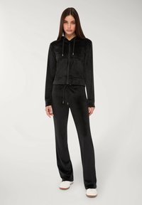 Tuta in velluto nero con giacca con cappuccio e zip sul davanti e pantaloni con coulisse, dal texture morbido e dalla silhouette aderente.