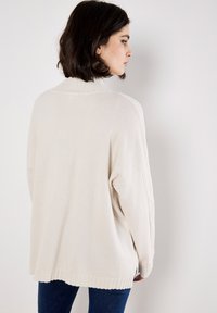 Kremowy, oversize'owy sweter z dzianiny z wysokim, prążkowanym dekoltem i bocznymi rozcięciami. Tkanina wydaje się być miękka o gładkiej fakturze.