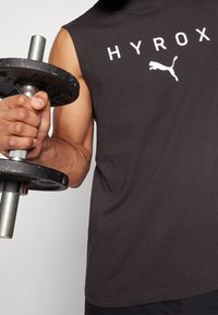 T-shirt de sport noir sans manches avec les logos "HYROX" et Puma en blanc. Le sujet tient un haltère en métal avec une prise texturée.