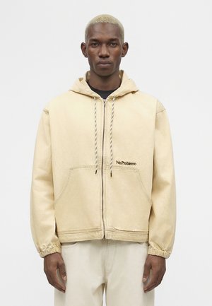 Uomo con capelli biondi corti che indossa una felpa beige con cerniera e scritta "No Problemo" e pantaloni chiari abbinati.