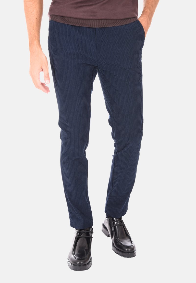 Uomo vestito con pantaloni di velluto a coste blu navy e scarpe in pelle nera con lacci, mentre sta in piedi con una mano in tasca su uno sfondo neutro.