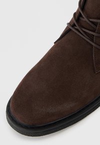 Rudas zomšos kulkšnies aulinukas su apvalia nosimi, tekstūruota paviršiaus ir juodu guminio pado. Viršuje yra susikryžiavusiu raišteliu dekoruota detalė.