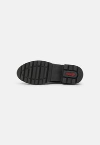 Semelle de chaussure en caoutchouc noir avec un motif texturé, présentant des rainures profondes et un logo rouge. Le design est plat et robuste pour une meilleure adhérence.