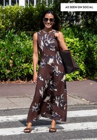 Donna sorridente che indossa occhiali da sole e un vestito lungo marrone a fiori, con una grande borsa a tracolla, mentre attraversa un passaggio pedonale.