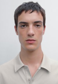 Ljusbeige polo-shirt med krage och knäppning med tre knappar, tillverkad av mjukt tyg, bärs av en person med kort mörkt hår, neutral bakgrund.
