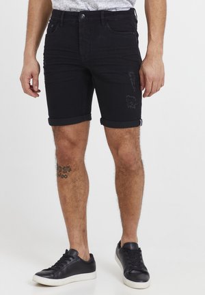 Sorte denim shorts med opfoldede manchetter, med slidte detaljer. Sammen med sorte sneakers og vist mod en hvid baggrund.