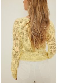 Pull jaune en maille avec un design en filet lâche, manches longues et ourlet ajusté. Le modèle porte un pantalon blanc, montrant une vue arrière texturée.