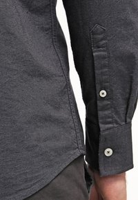 Dunkelgraues, strukturiertes Langarmshirt mit spitzem Kragen und geknöpften Manschetten. Mit silbernen Knöpfen und taillierten Details.