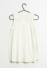 Crèmekleurige mouwloze top hangend aan een zwarte metalen hanger. Voorzien van een geribbelde halslijn en zoom, met een gladde, lichtgewicht textuur.