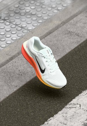 Chaussure de course Nike blanche avec swoosh noir et semelle dégradée orange sur l’asphalte près d’un pavé tactile et d’un passage piéton.